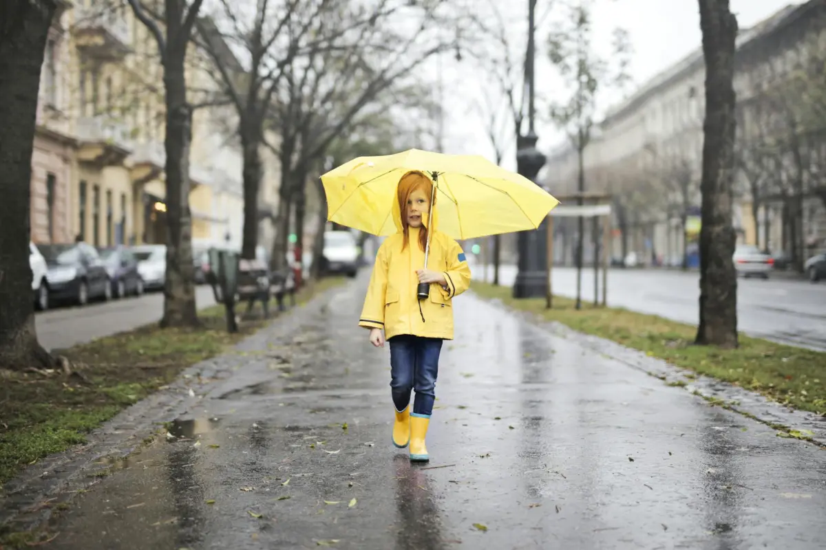 A child wearing a yellow raincoat walks with an umbrella on a wet city street.