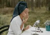 A woman in a bathrobe applies face cream in a serene outdoor environment.