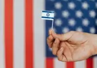 Closeup of crop unrecognizable person holding small flag of Israel before huge flag of United States of America on background