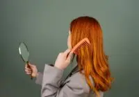 Red-haired woman using a comb while holding a mirror, focusing on hair care and beauty.