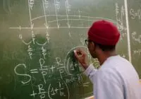 Student writing complex mathematical equations on a chalkboard in a classroom setting.