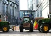 Tractors block a city street in a protest near modern buildings, waving a Belgian flag.