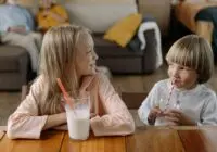 Two children smiling and drinking milk with straws indoors, enjoying each other's company.