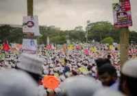 A large crowd gathers outdoors with vibrant banners and signs, showcasing a cultural event or demonstration.