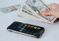 Calculator application on mobile phone screen on white surface near crop anonymous person holding pile of dollars
