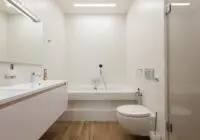 Interior of modern light bathroom with white bathtub and cabinet in front of shower cabin with glass doors