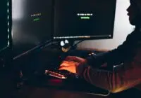 Side view of crop anonymous male cyber thief accessing information on desktop computer screens at dusk