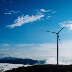 जलवायु संकट भारत की भूमिका: वैश्विक प्रयासों में अग्रणी कदम 24 A solitary wind turbine rises against a vibrant blue sky, surrounded by clouds and distant mountains.