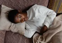 An adult woman relaxing indoors with artistic face paint, lying on a pillow and blanket in a cozy setting.